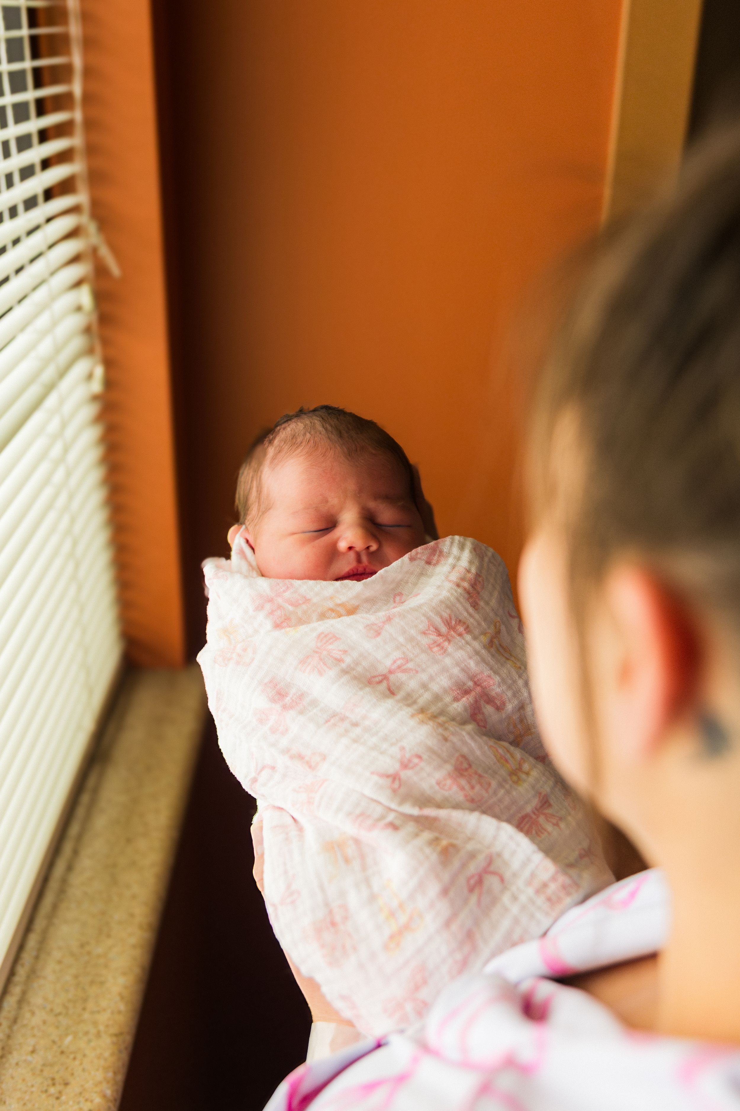 Newborn baby swaddled in white muslin blanket being held by mother near sunny window.