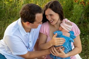 A couple embraces while holding their newborn baby in a sunny meadow with wildflowers.