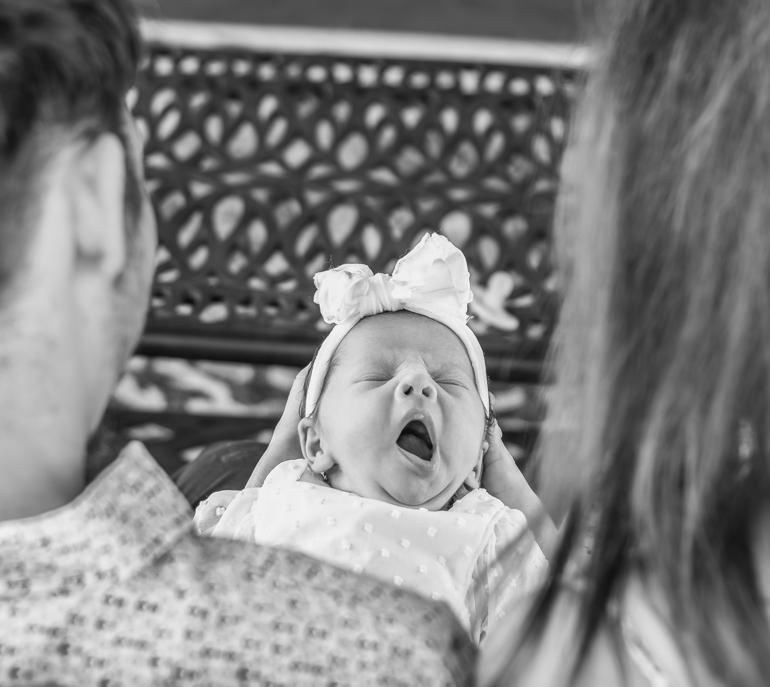 Baby yawns while being held by parents on a park bench.