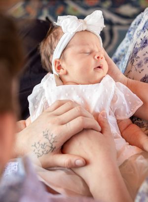 Sleeping newborn in white dress and bow held by caring hands.