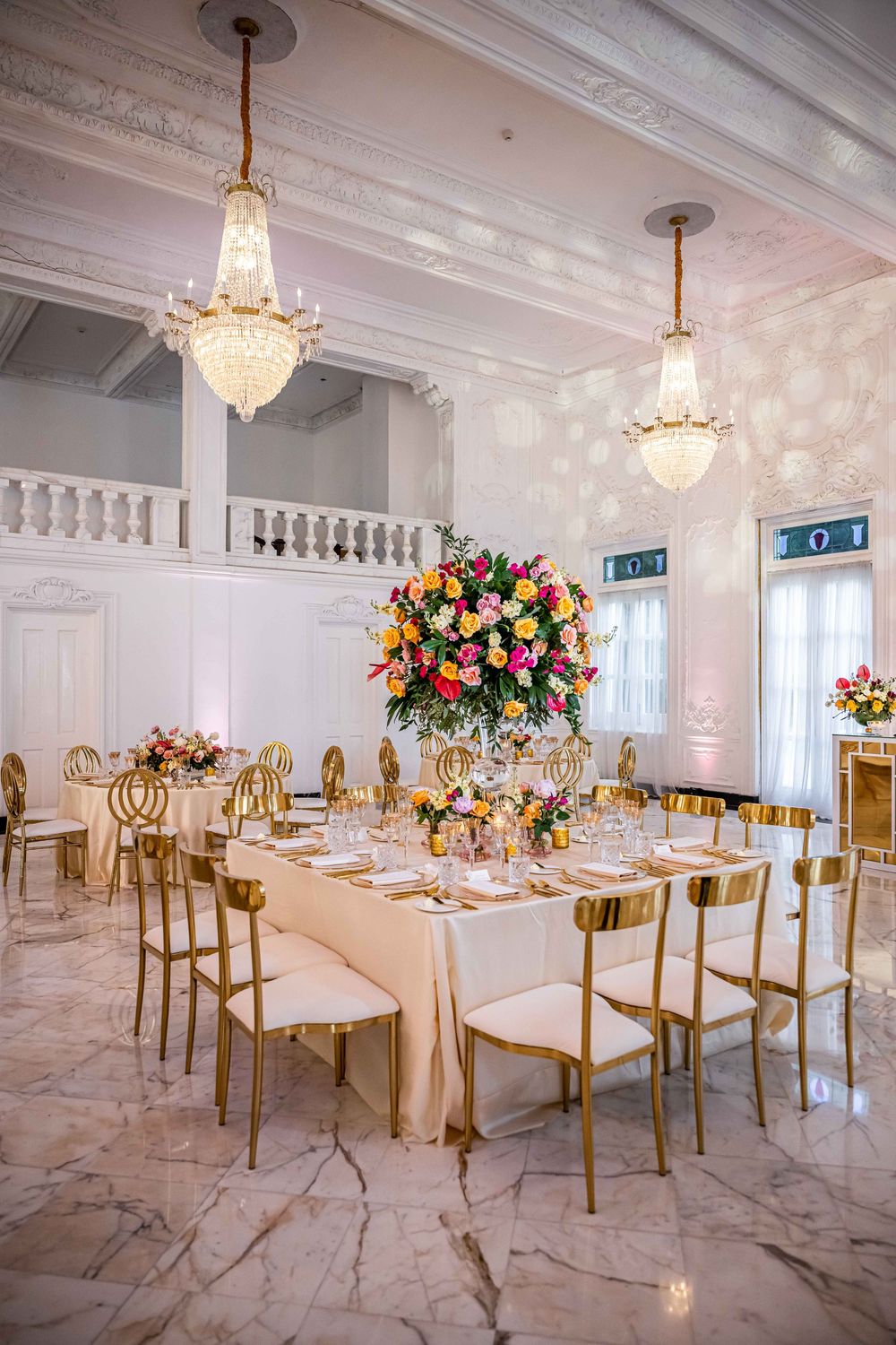 Elegant wedding reception table with gold chairs, crystal chandeliers and dramatic floral centerpiece in white marble ballroom.