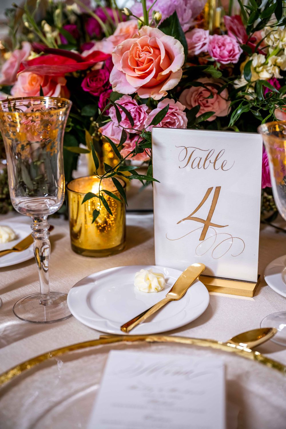 Elegant wedding table setting with pink and red floral centerpiece, gold cutlery, and Table 4 card on white linens.
