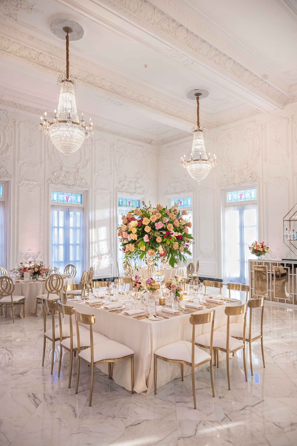 Elegant wedding reception table with crystal chandeliers and pink floral centerpiece in luxurious white marble ballroom.