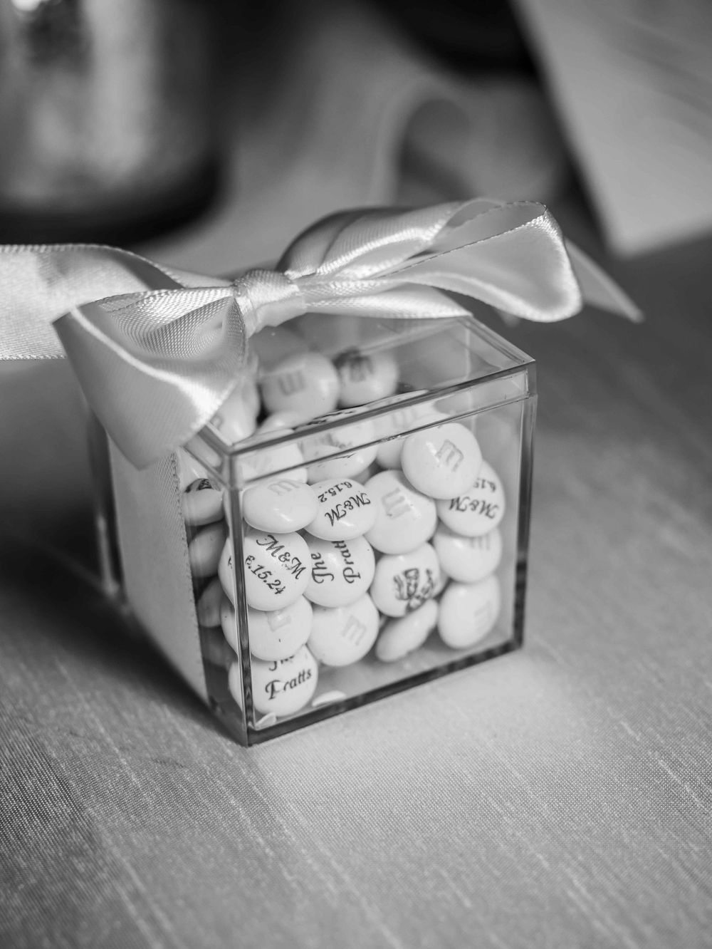 Clear gift box filled with personalized candies and tied with a white satin bow on a gray surface in black and white.