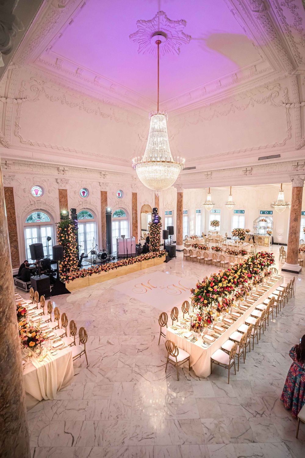 Elegant ballroom setup with long white dining tables and crystal chandelier under ornate ceiling with Christmas trees.