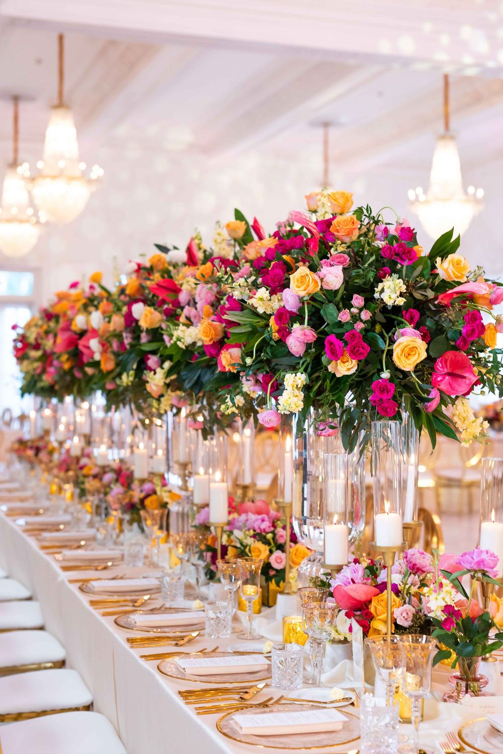 Vibrant floral centerpieces with pink, orange and yellow blooms line a formal white dining table with gold accents.