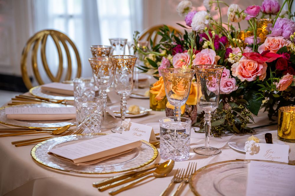 Luxurious wedding table setting with pink roses, crystal glassware, gold flatware and white table linens.