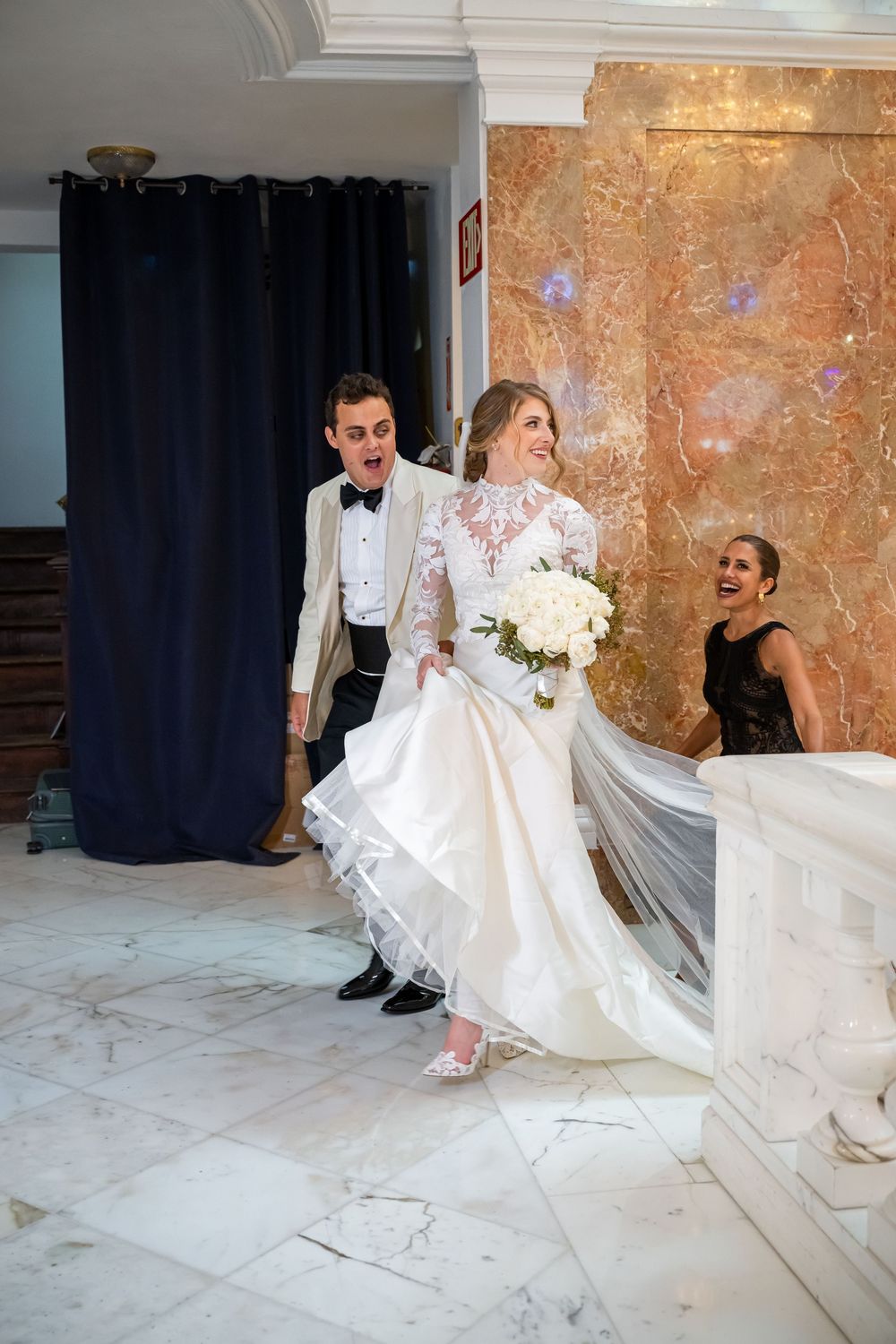 A bride in a white lace gown with long sleeves walks up marble steps with her wedding party in formal attire.