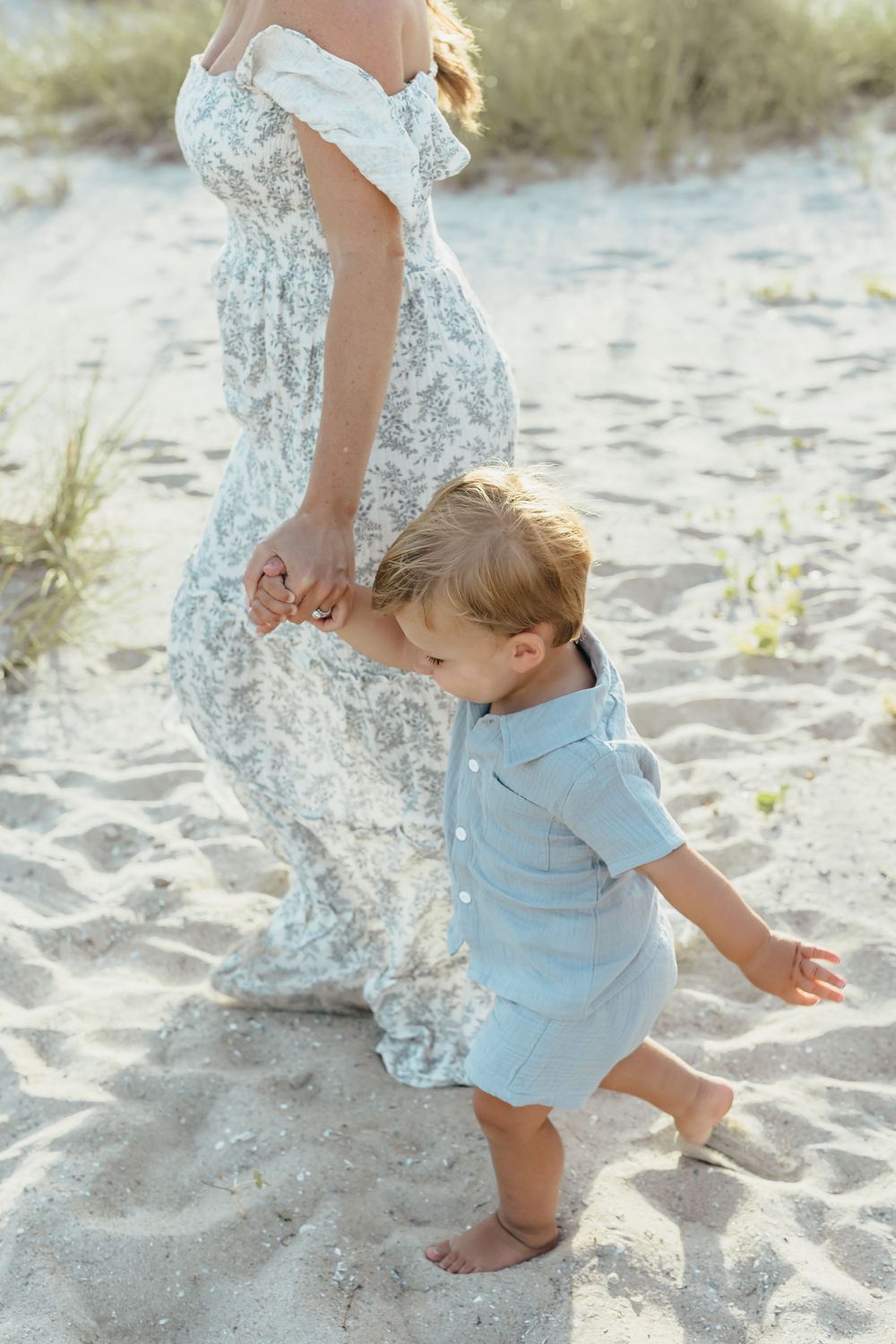 A toddler in a blue romper holds his pregnant mother's hand on a sandy beach.