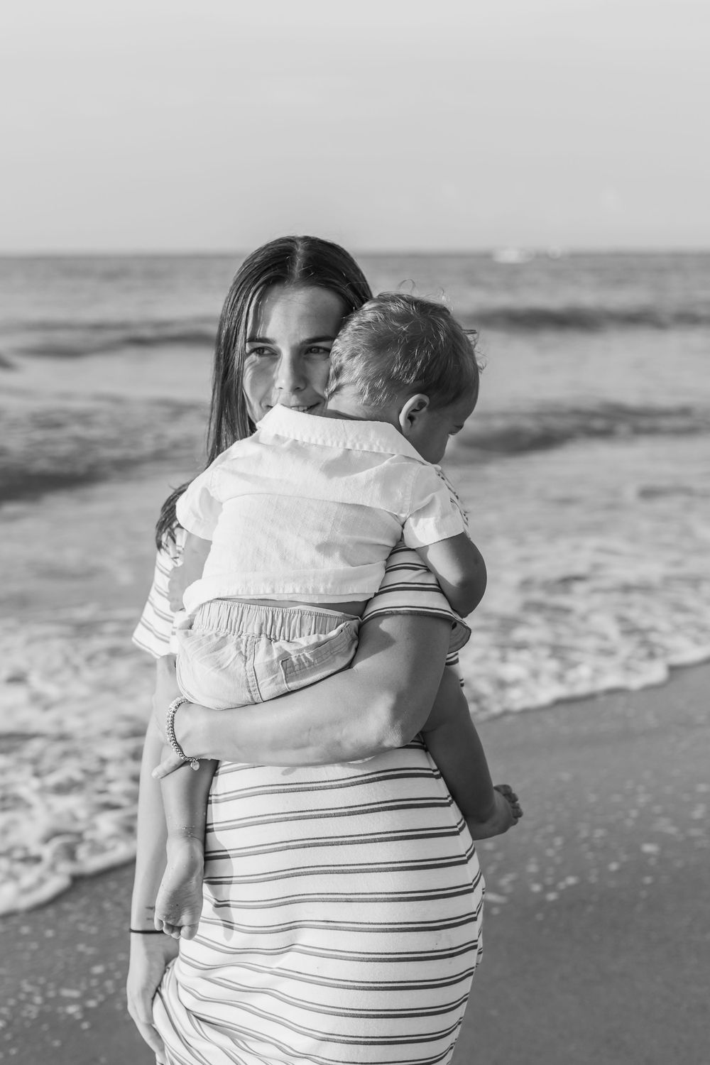 Black and white photo of young mother holding baby on sandy ocean beach.