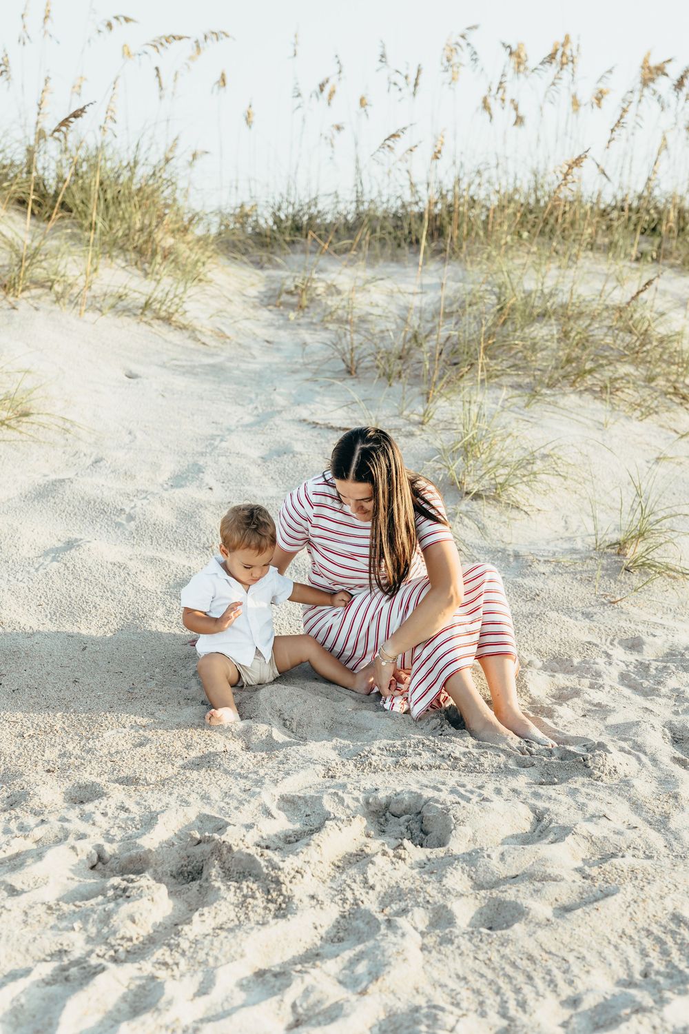 Mother in striped dress sits with toddler on sandy beach dunes with sea oats in background.