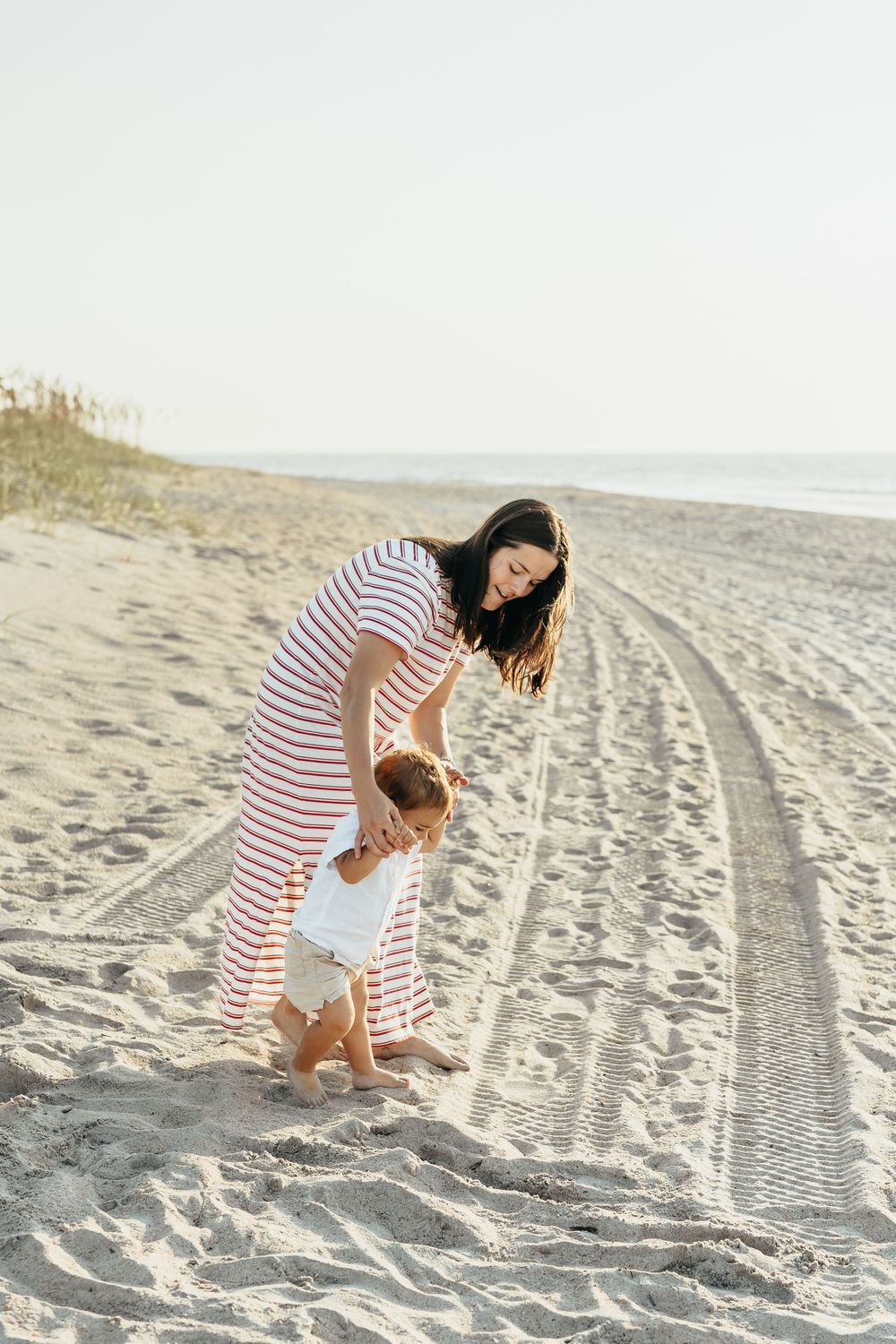 Young mother in striped dress helping toddler walk on sandy beach with ocean in background.