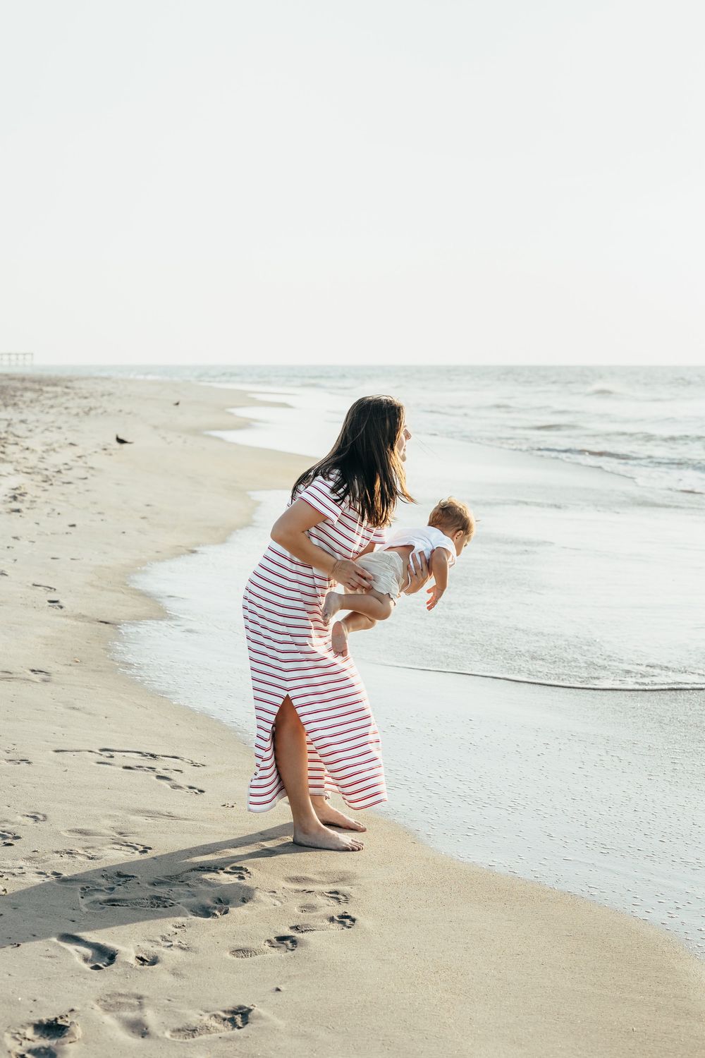 A woman in a striped dress holds a baby while standing at the edge of the ocean on a sandy beach.