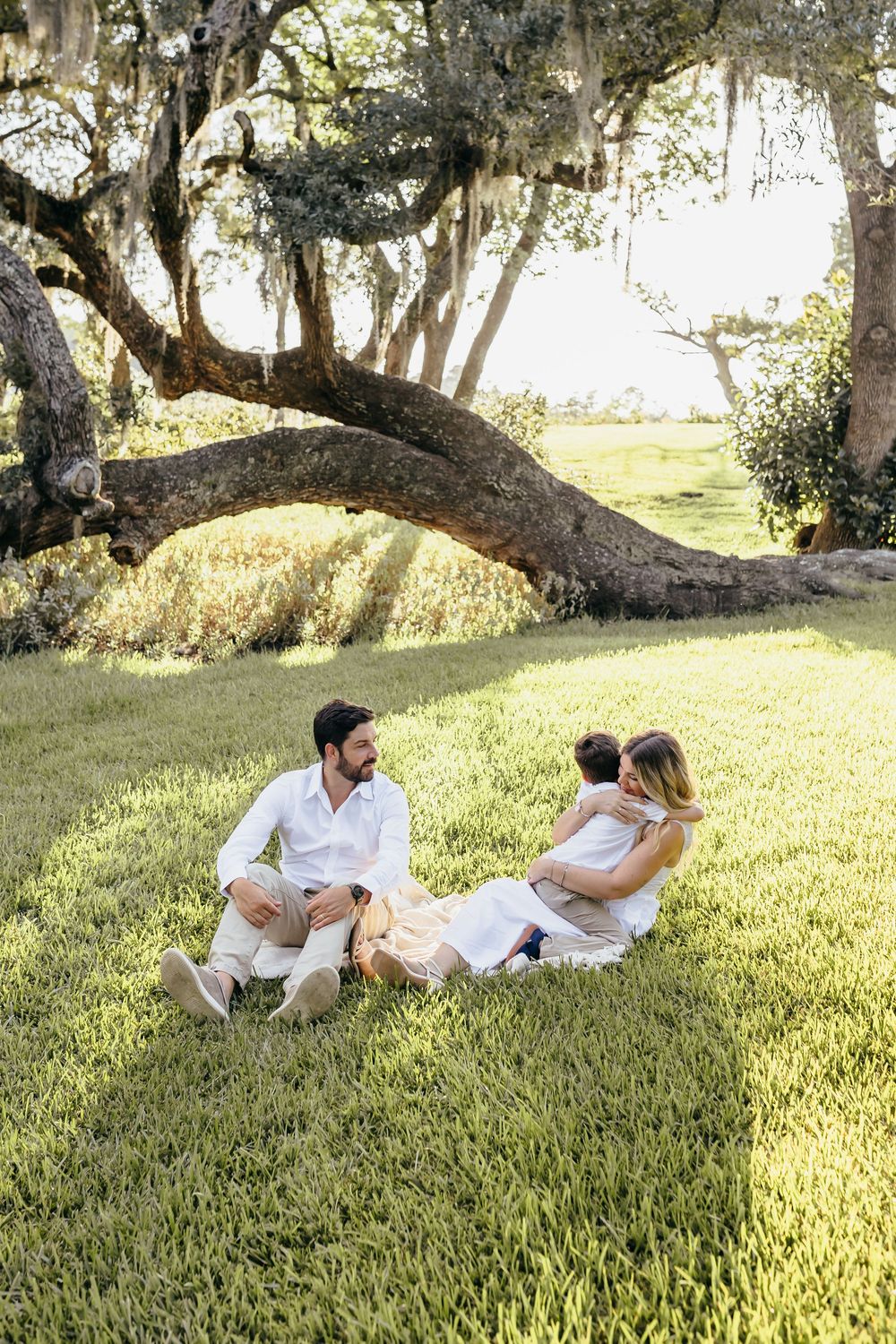 Family sitting on lush green grass under a sprawling oak tree with Spanish moss, enjoying a sunny outdoor moment.