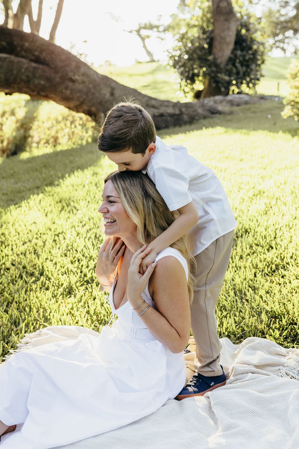 A smiling woman in white sits on a blanket outdoors as a young boy hugs her from behind near a tree.