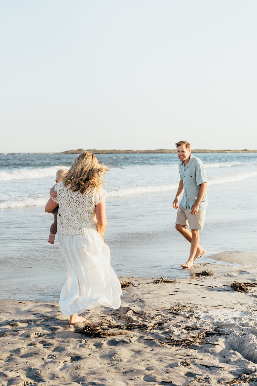 Family of three enjoying a sunny beach walk, mother in white dress holding baby while father approaches barefoot on sandy shore.