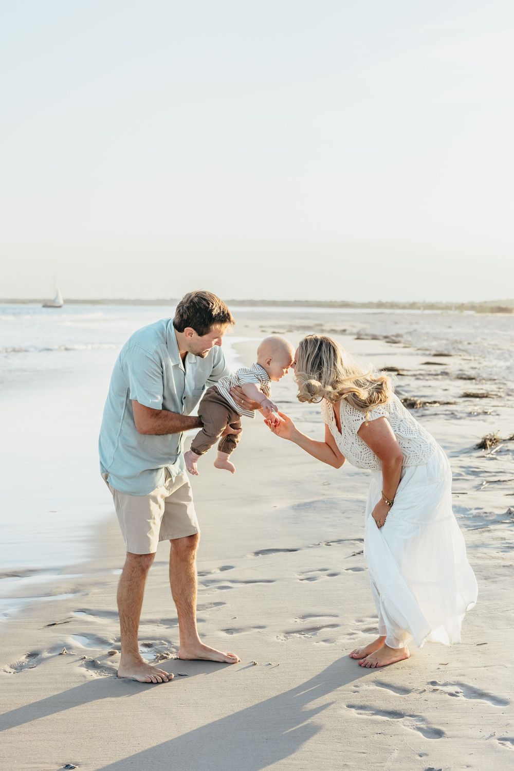 A young family plays with their baby on a sunny beach, parents in casual attire sharing a joyful moment.
