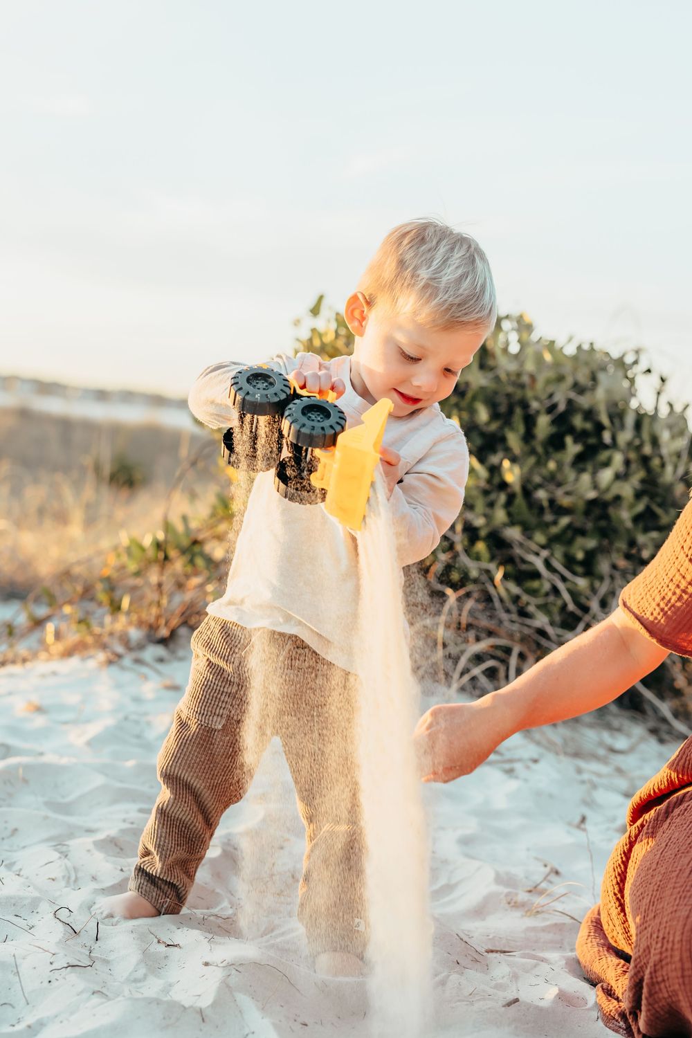 Toddler playing with toy trucks in white sand on a beach, pouring sand with an adult nearby.