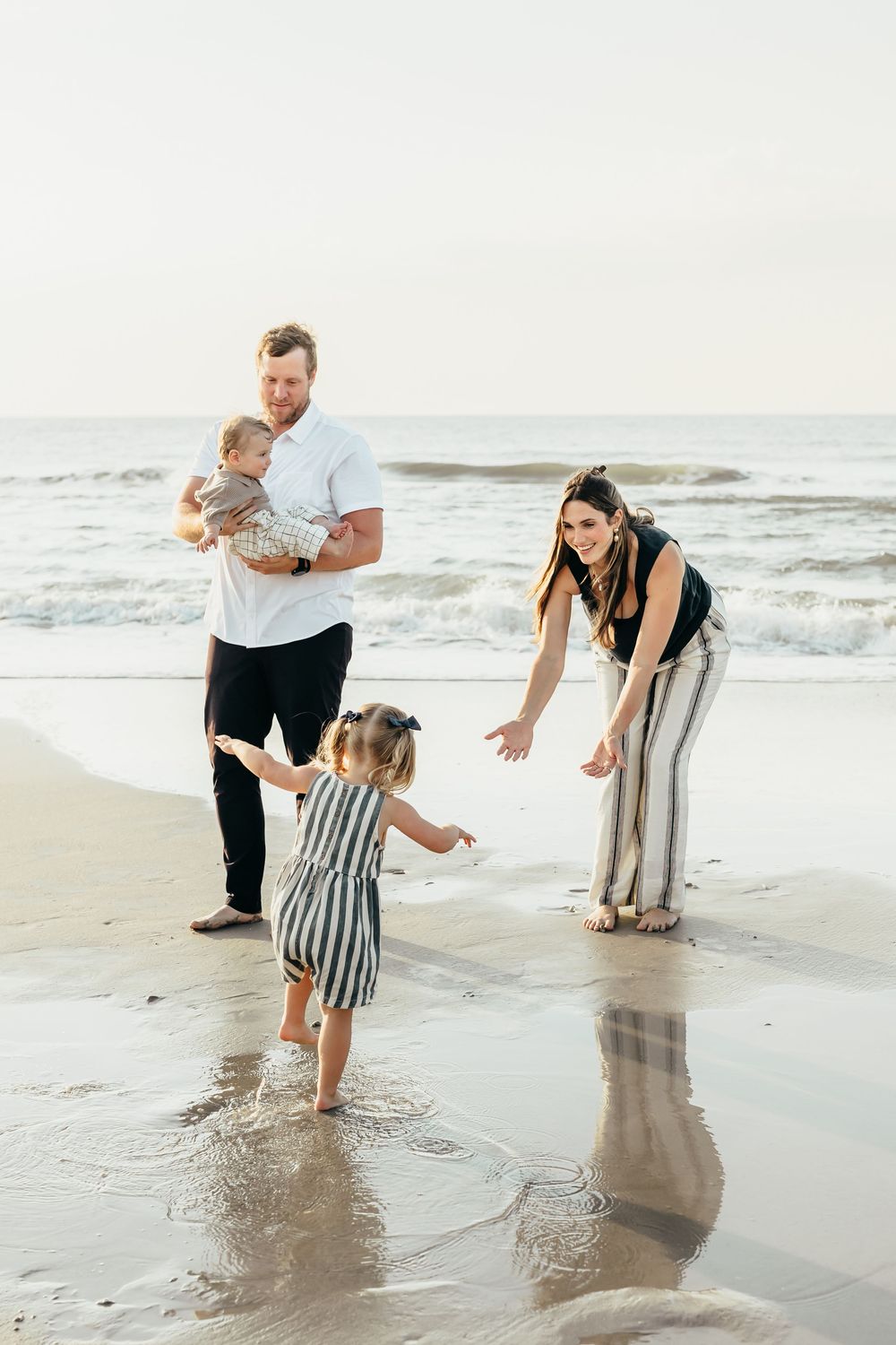 Family of four enjoying a sunny beach day, with parents playing with toddler girl and baby by ocean waves.