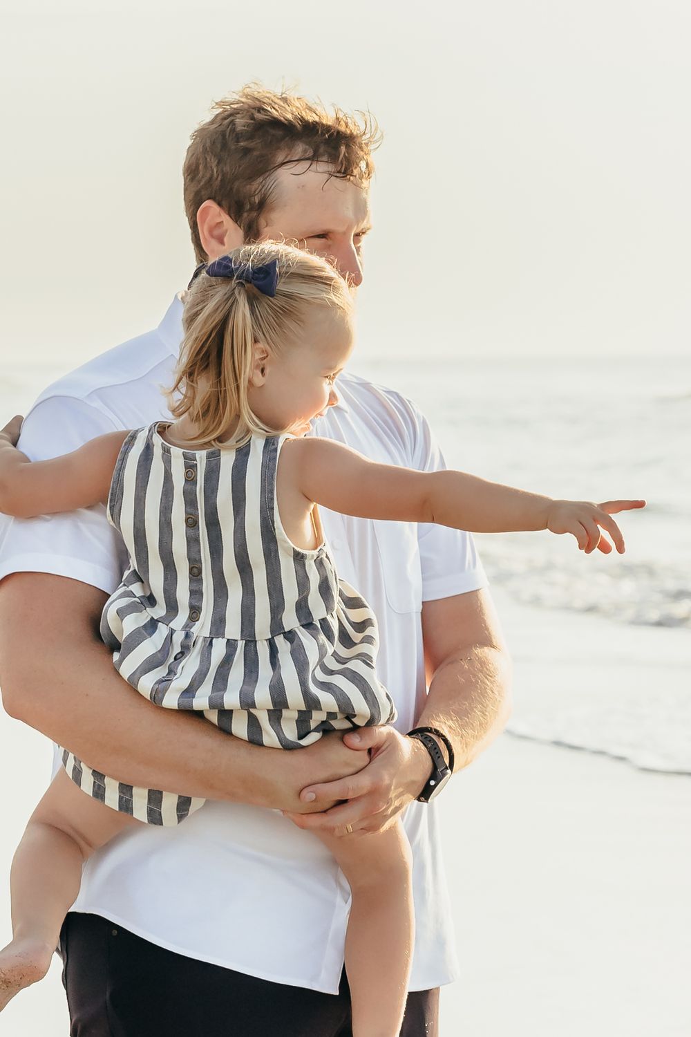 Father holding young daughter on a sunny beach, as she points into the distance near ocean waves.