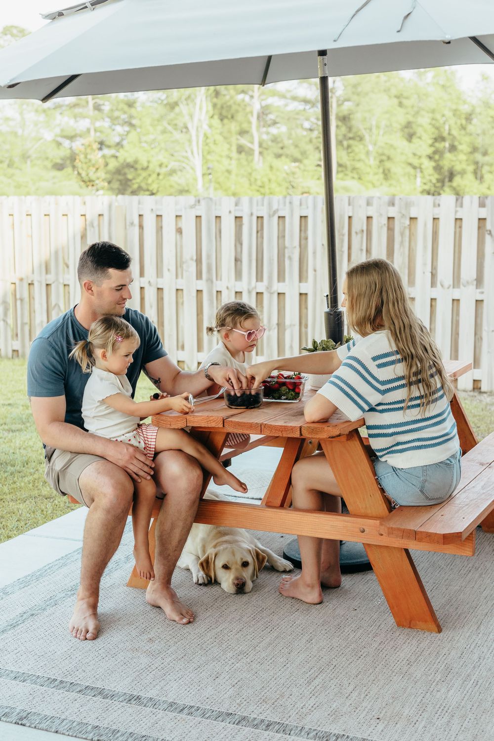 Family enjoying outdoor picnic at wooden table with umbrella, dog resting underneath, in fenced backyard.