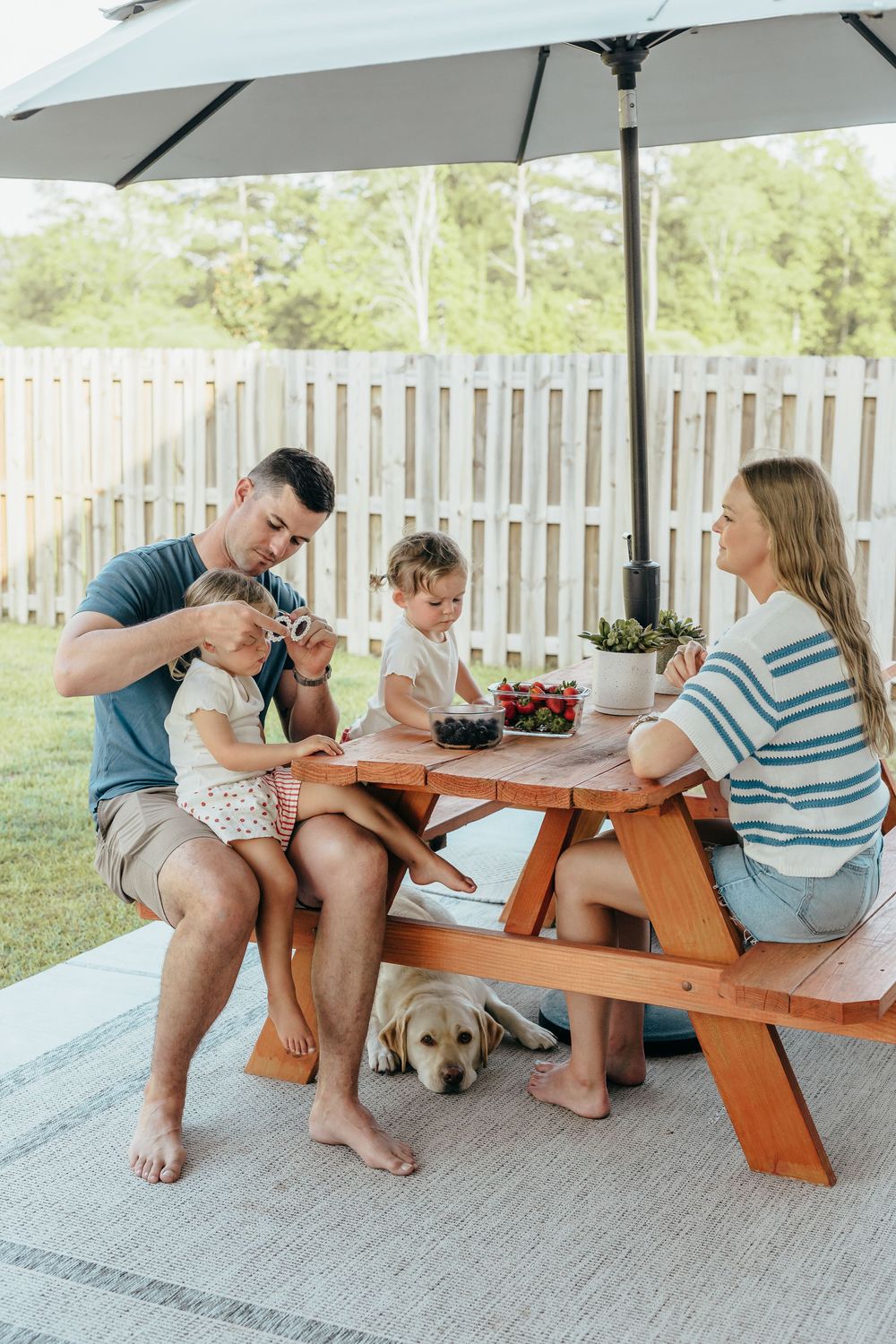 Family enjoying outdoor picnic at wooden table with umbrella, two toddlers, Labrador dog, and fresh strawberries in backyard.