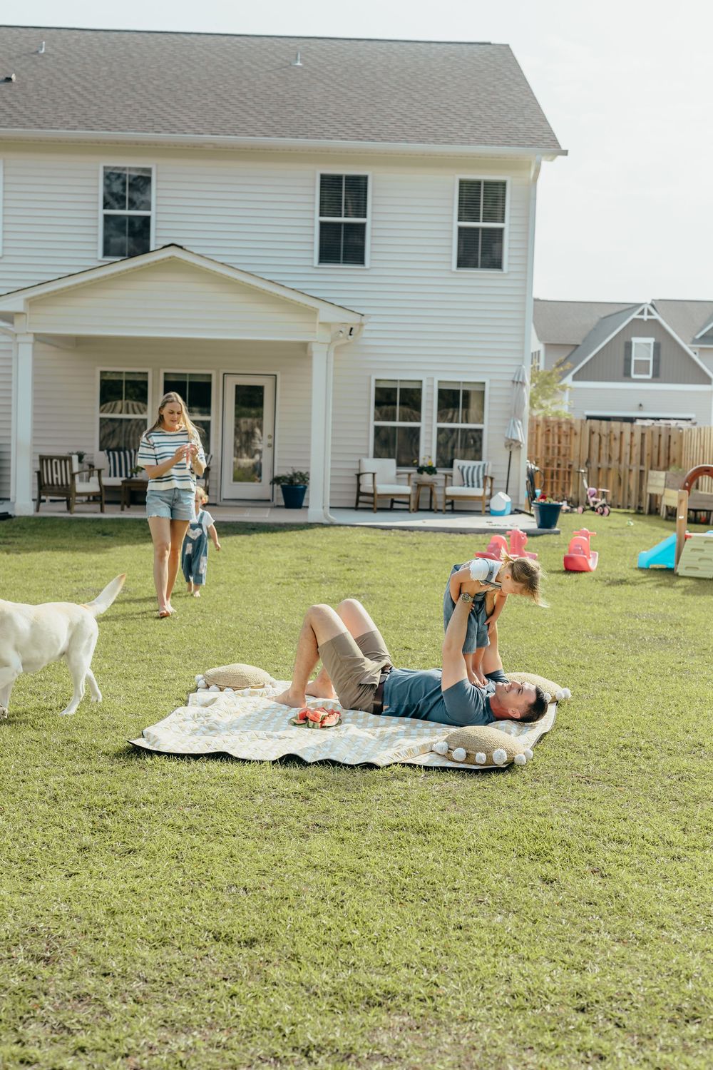 Family enjoying a sunny backyard day with dad playing on blanket, mom holding baby, and white dog on green lawn.