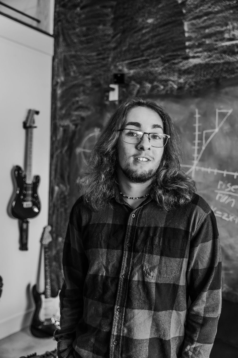 Black and white portrait of someone with long hair and glasses standing against a chalkboard wall with a guitar mounted nearby.