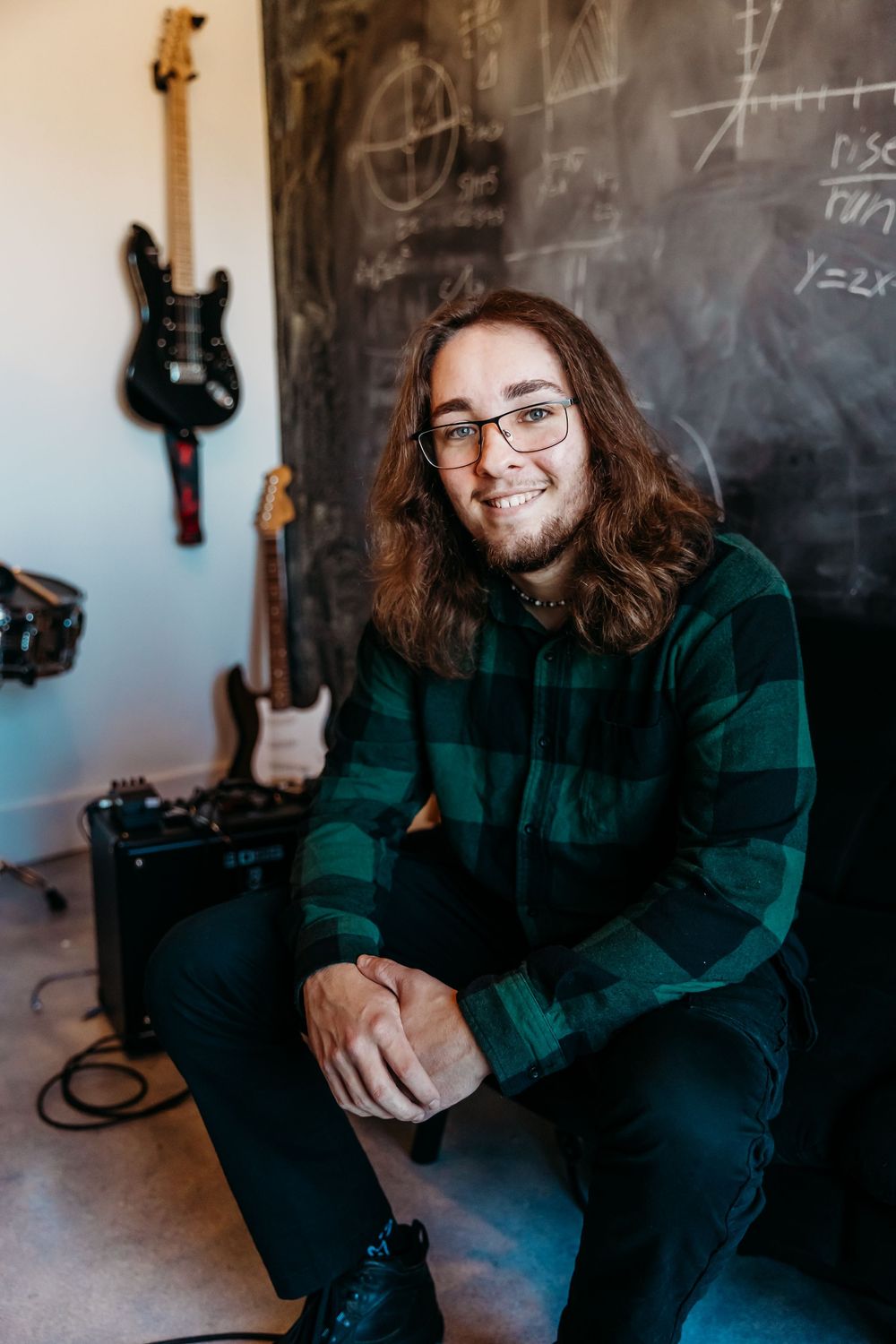 In a music room with a chalkboard and guitars, someone wearing a green plaid flannel shirt and glasses sits casually smiling.