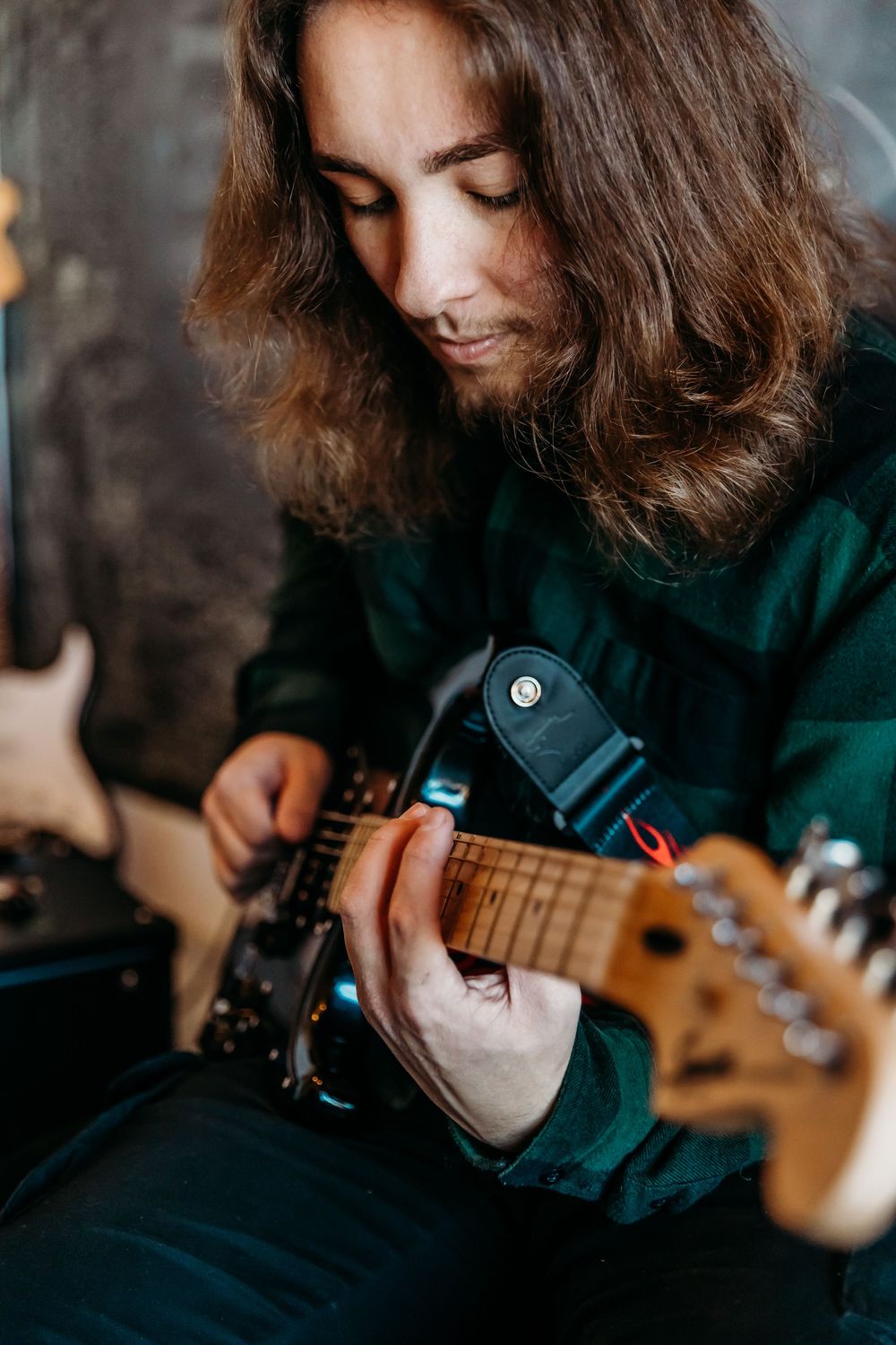 Musician with long hair wearing a green hoodie plays an electric guitar, shown in closeup profile view.