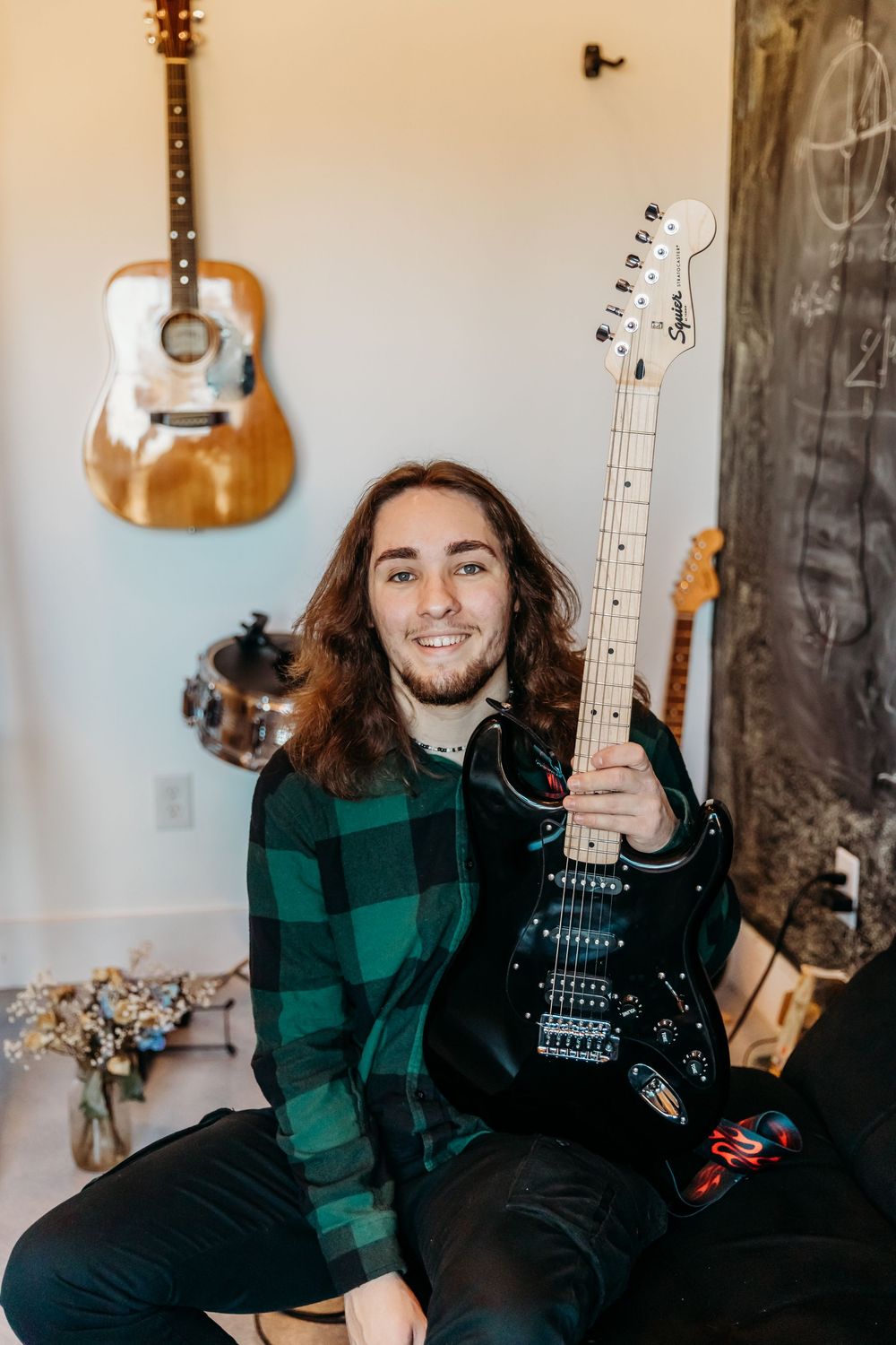 Musician in green plaid flannel holding black electric guitar with acoustic guitar mounted on wall in background.