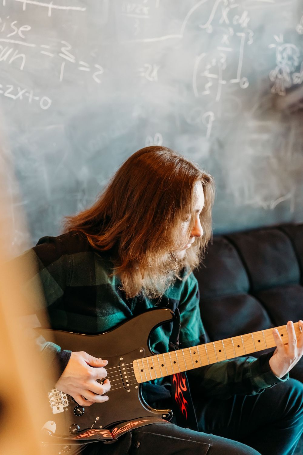 A musician with long hair plays electric guitar against a chalkboard backdrop in casual attire.