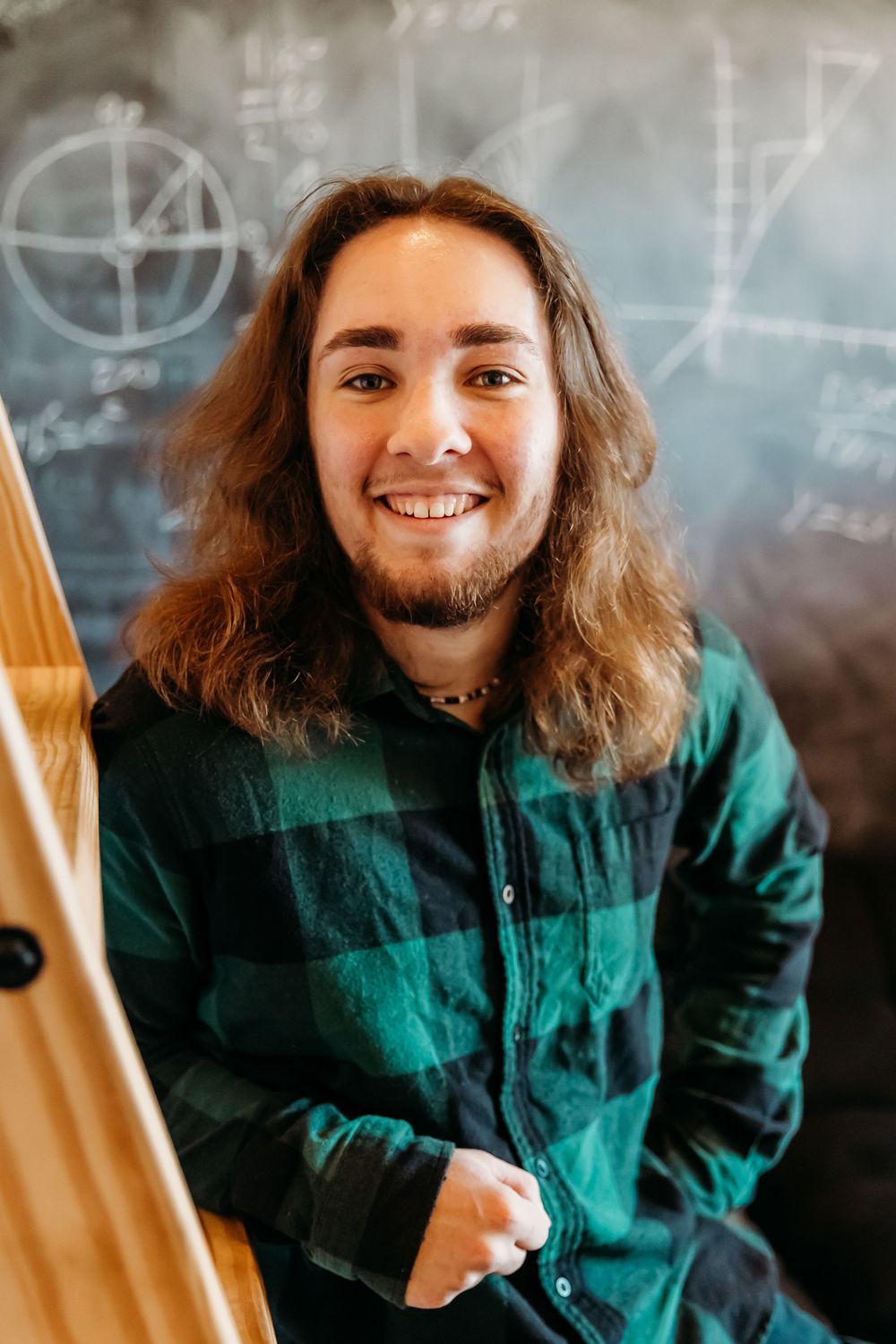 Portrait of someone with long wavy brown hair wearing a green plaid flannel jacket and smiling in front of a chalkboard wall.