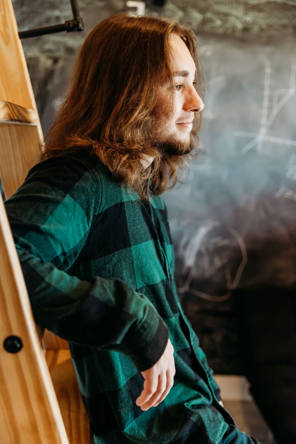 Person in green and black plaid flannel shirt poses in profile view against wooden wall with smoke in background.