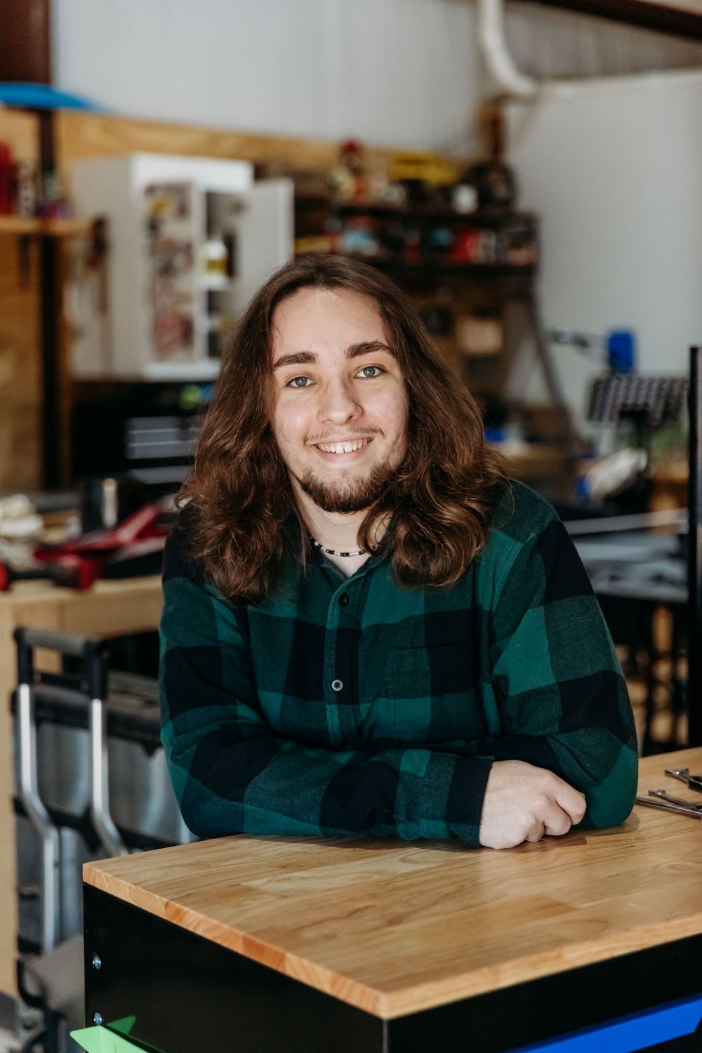 A person with long brown hair wearing a green plaid flannel shirt sits at a wooden desk in a workshop environment.