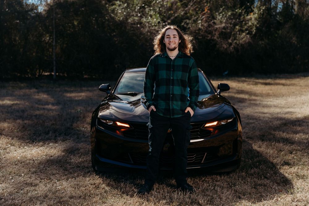 Someone in a plaid shirt poses confidently in front of a black sports car with glowing headlights at sunset.