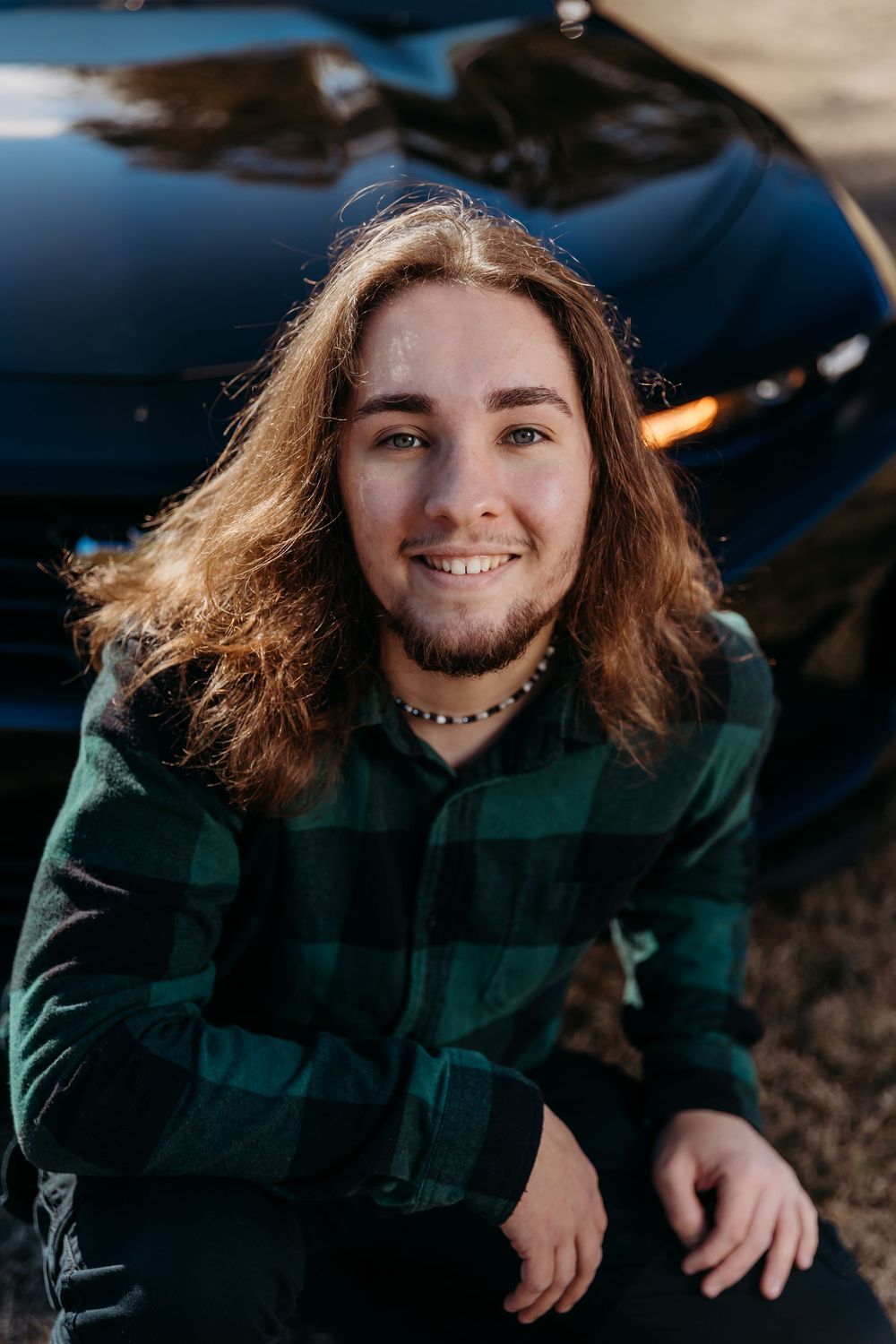 A person in a green plaid flannel jacket poses outdoors with long brown hair and a necklace.