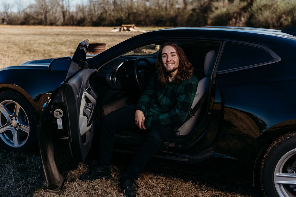 Black luxury sports car parked on dirt road with open driver door during daytime in rural setting.