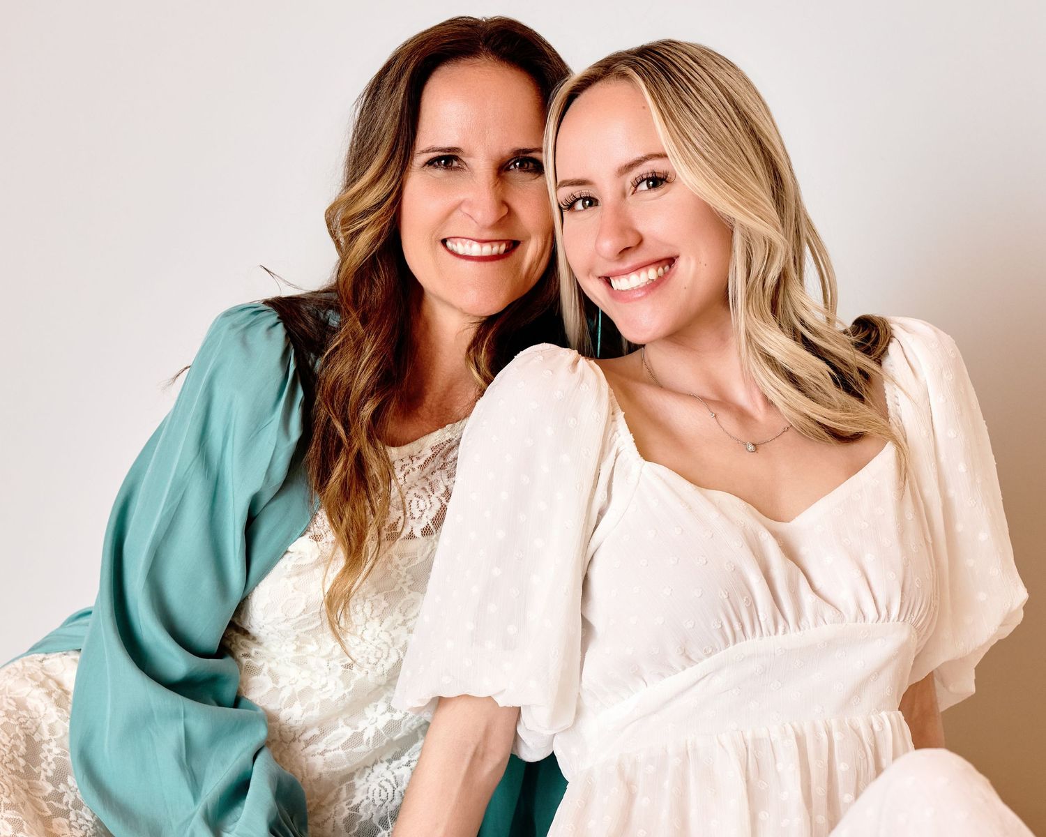 Joyful studio portrait of a smiling mother with brown hair and daughter with blonde hair, posing closely. Professional family photography.