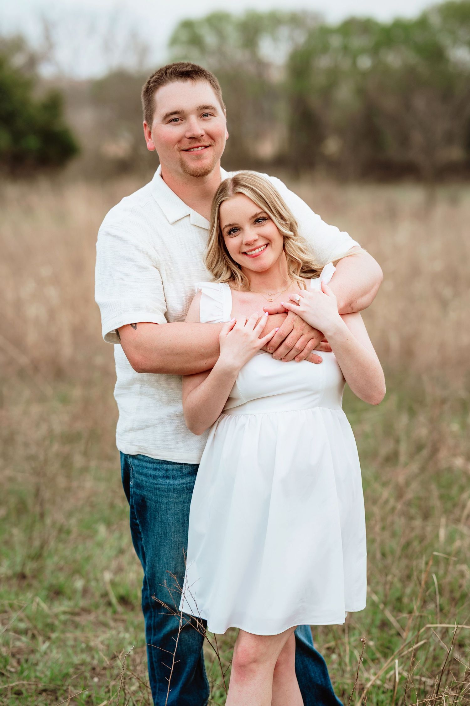 Romantic couple embraces in a spring field with blooming trees and greenery in the background.