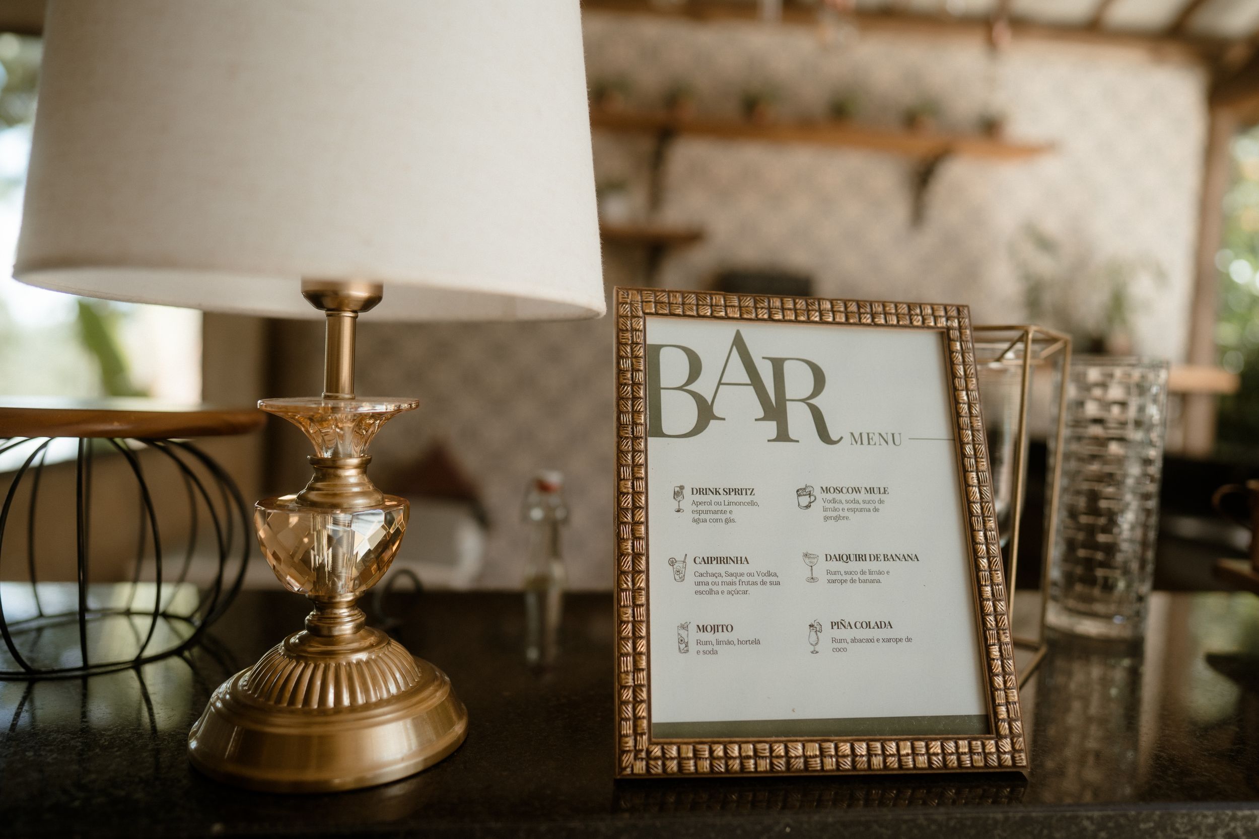 Gold-framed bar menu sign displayed next to an elegant table lamp on a dark surface.