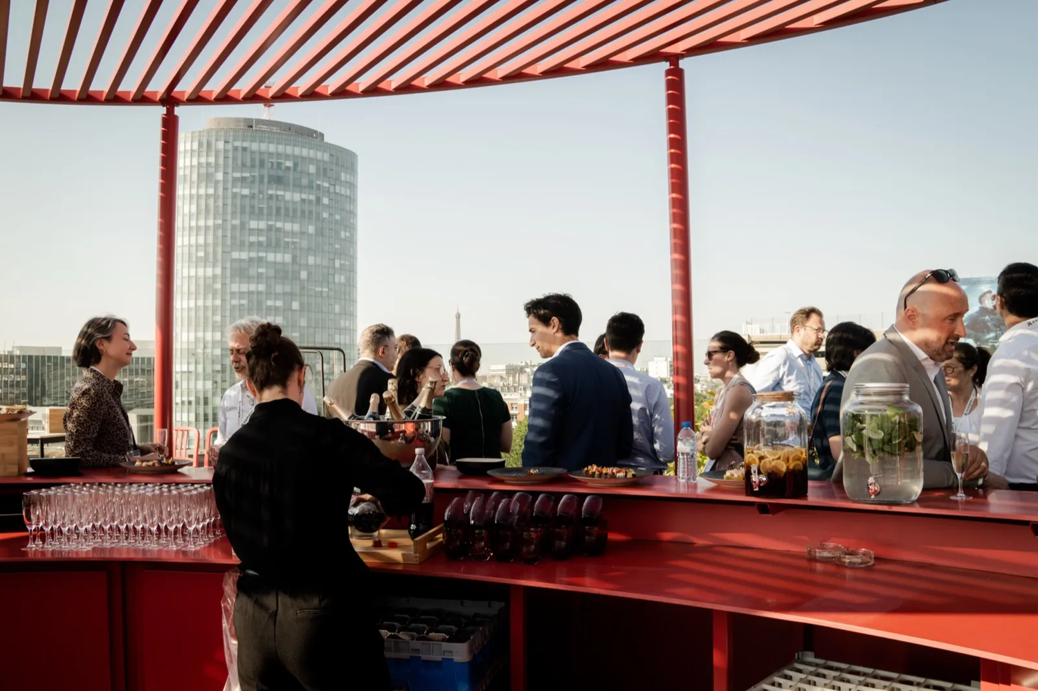 Groupe de personnes rassemblées autour d’un bar extérieur sous une structure rouge, avec une tour en arrière-plan.