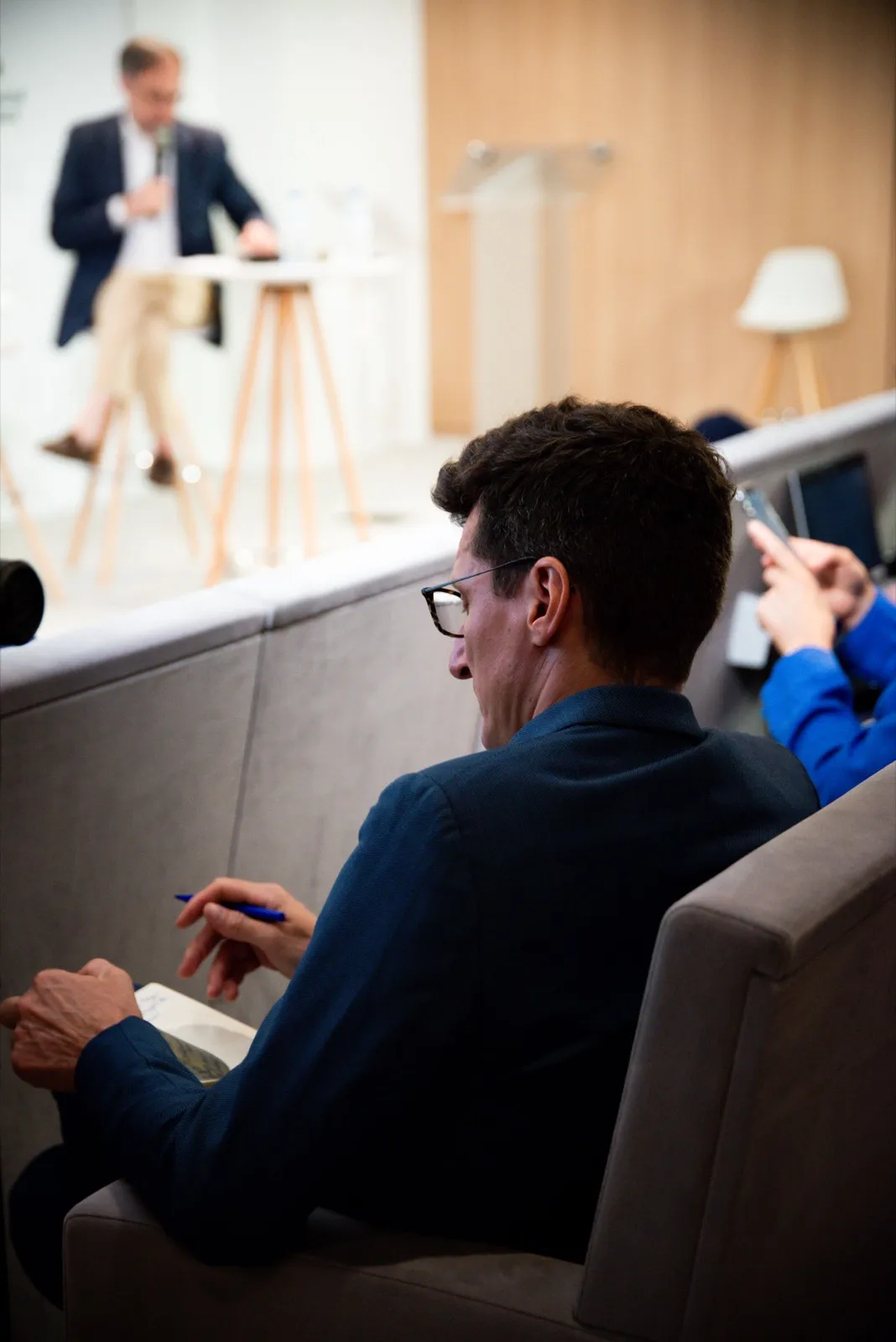 Homme concentré, installé confortablement avec un ordinateur portable, scène en arrière-plan avec un intervenant debout.