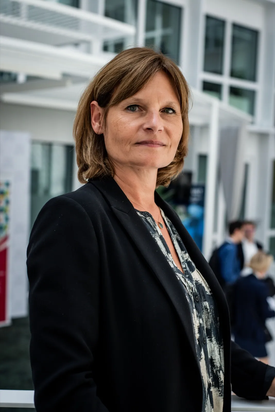 Femme souriante en blazer noir, regard vers l’objectif, fond de bureaux vitrés avec collègues flous.