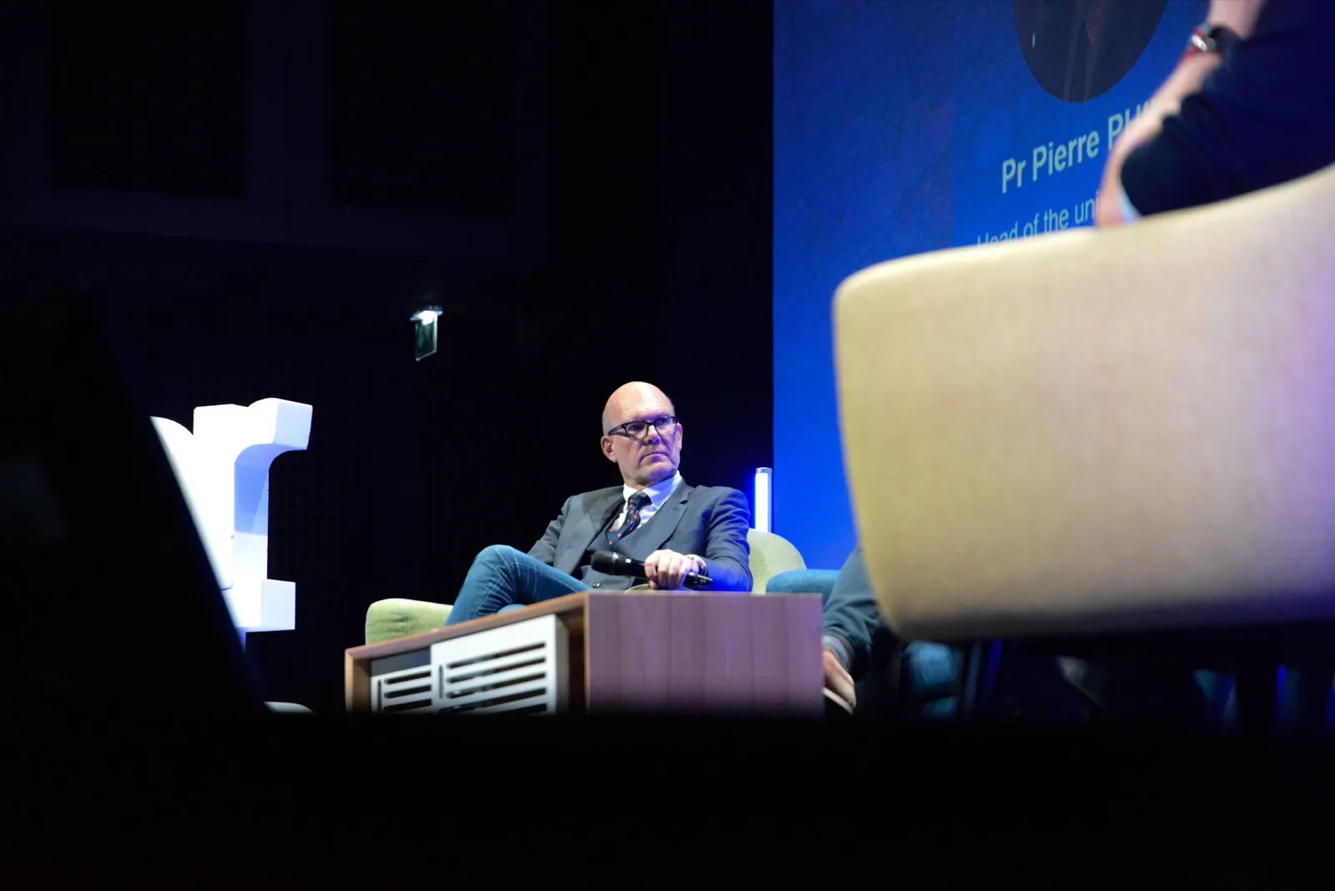 Homme assis dans un fauteuil sur scène, éclairé par un projecteur, participant à un panel professionnel.
