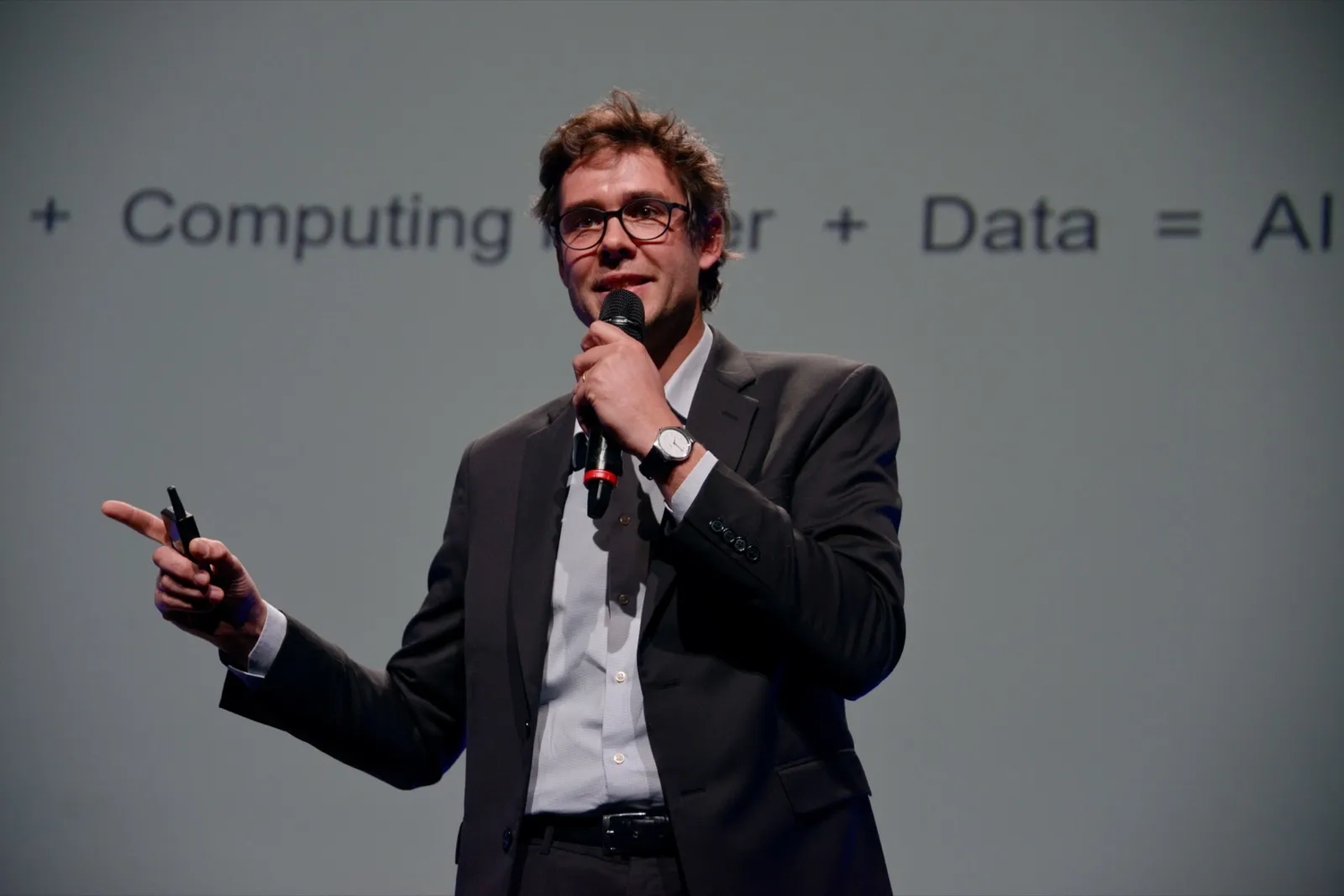 Homme en costume tenant un micro et une télécommande, en train de faire une présentation sur l’IA devant un écran de projection.