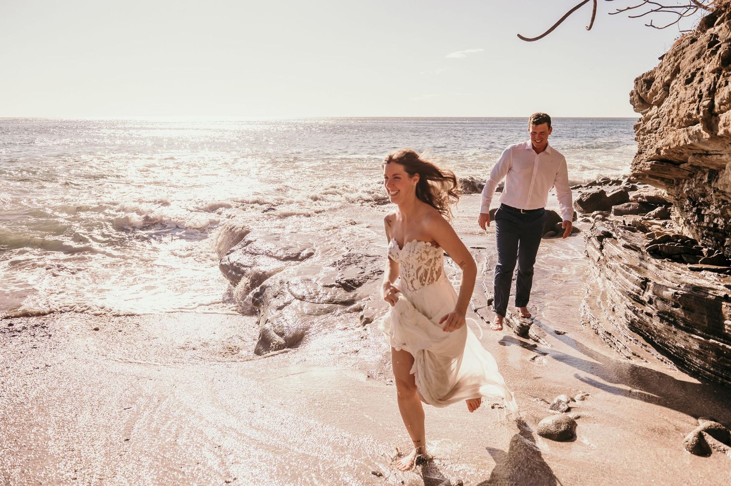 Two people in light flowy attire hold hands while walking along rocky coastal cliffs at sunset.