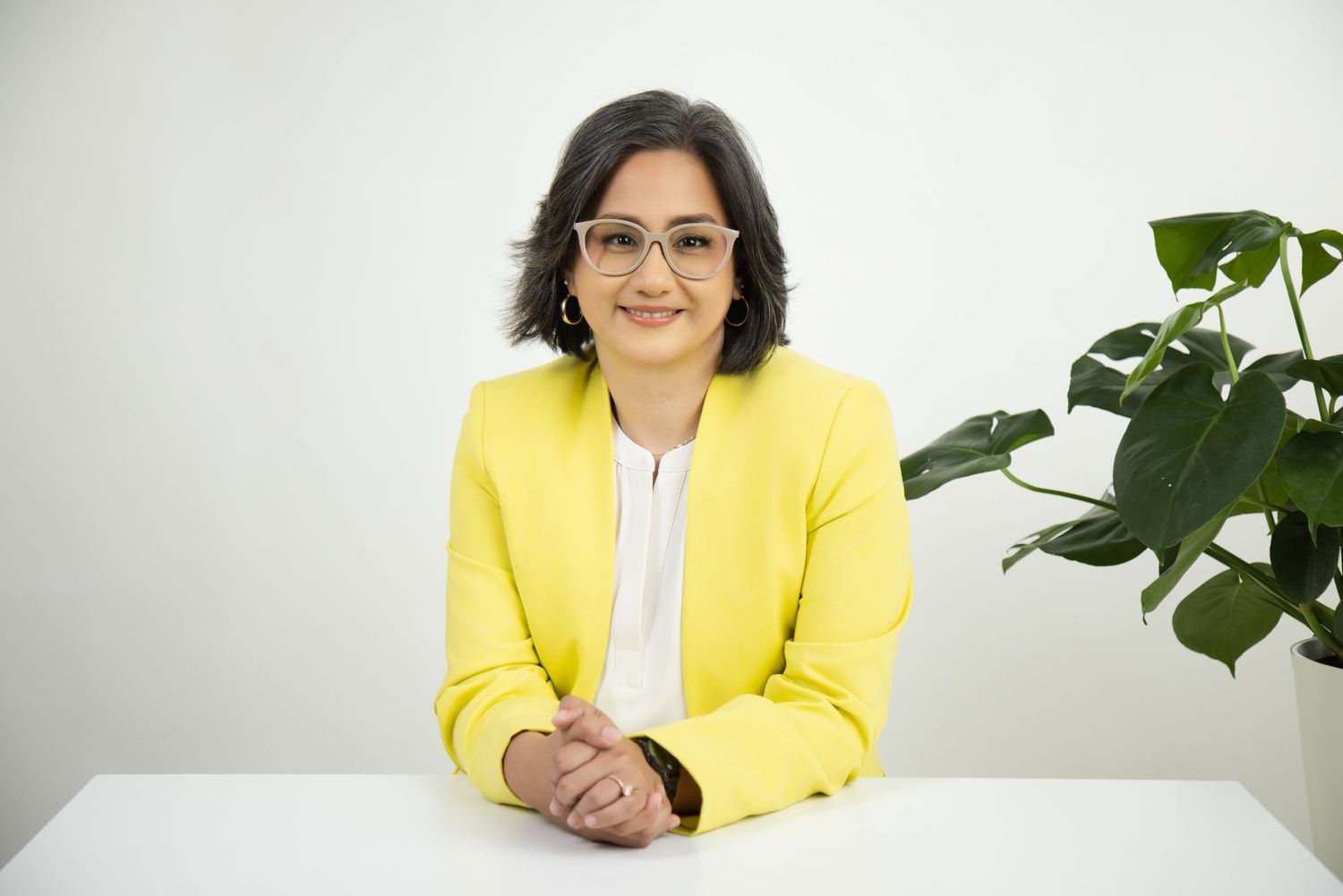 Professional in a bright yellow blazer and glasses sits at a white desk with a green plant in the background.