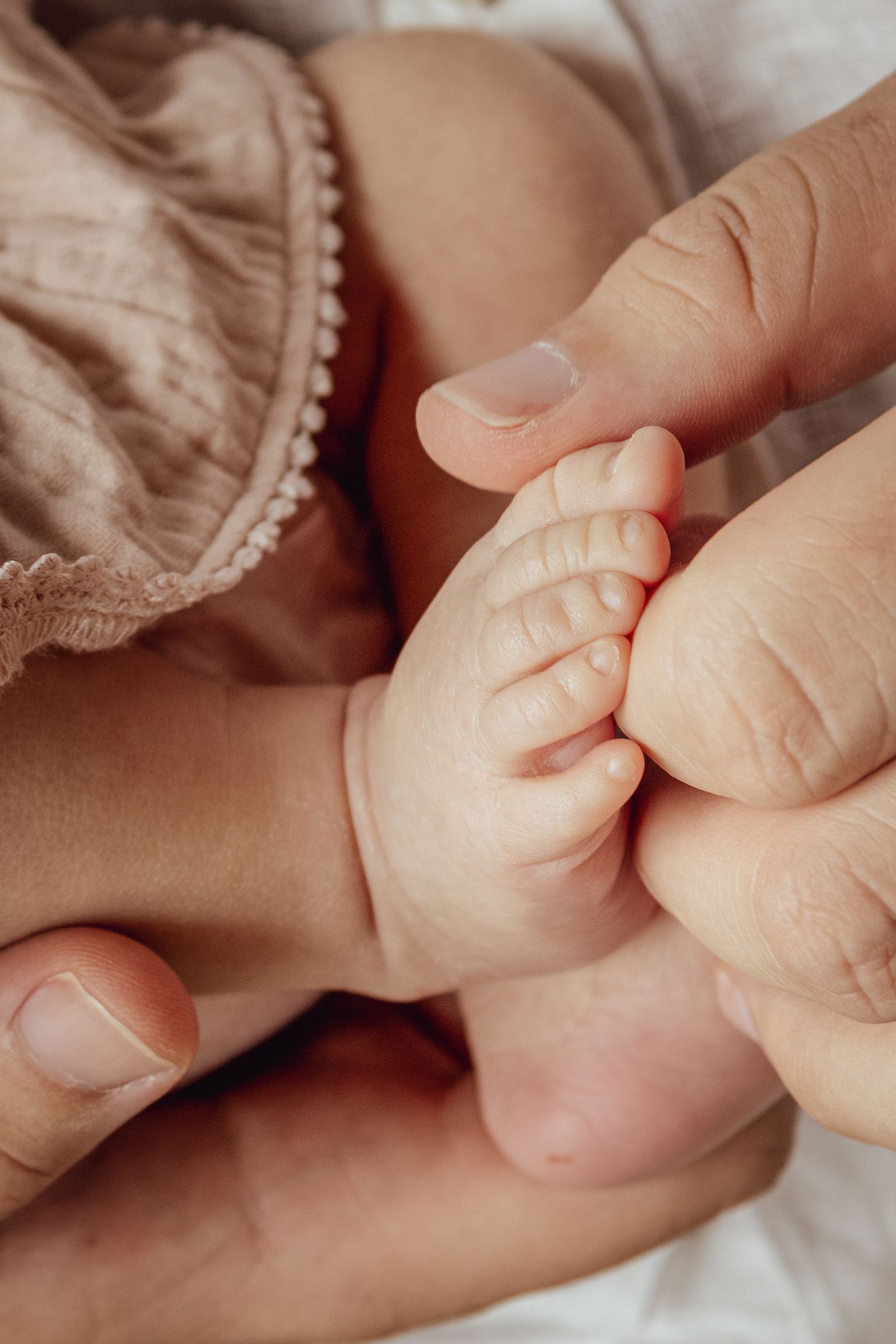 A tiny baby's foot being gently held by adult hands against a soft beige fabric background.