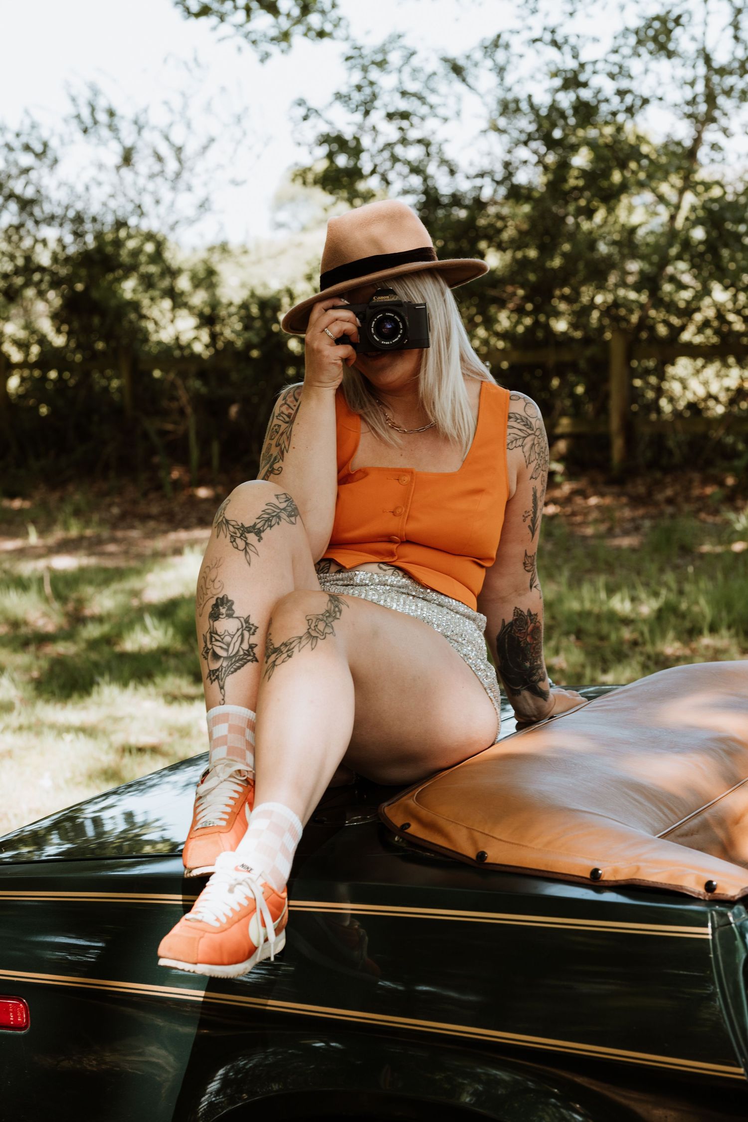 A photographer in an orange top and denim shorts sits on a vintage car taking pictures with a camera in summer.